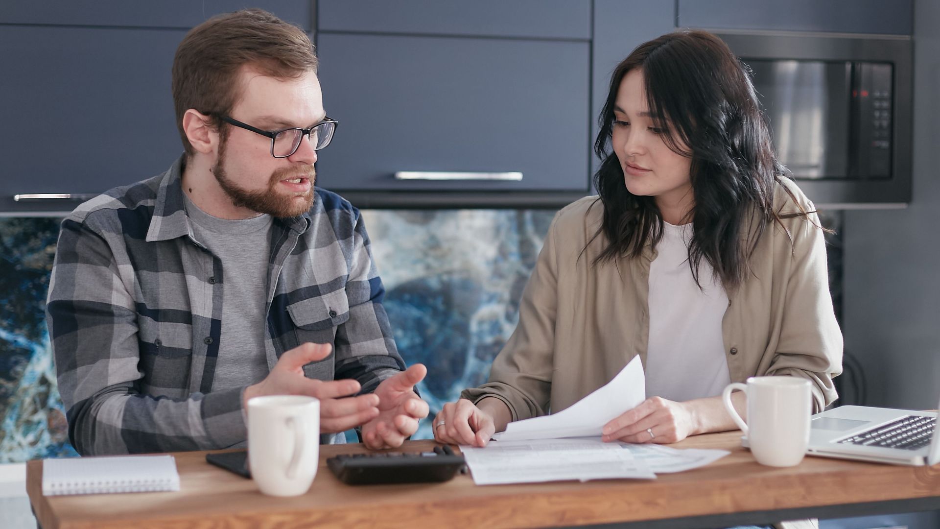 A couple engaged in a financial discussion, sitting at a kitchen table with papers and a laptop.