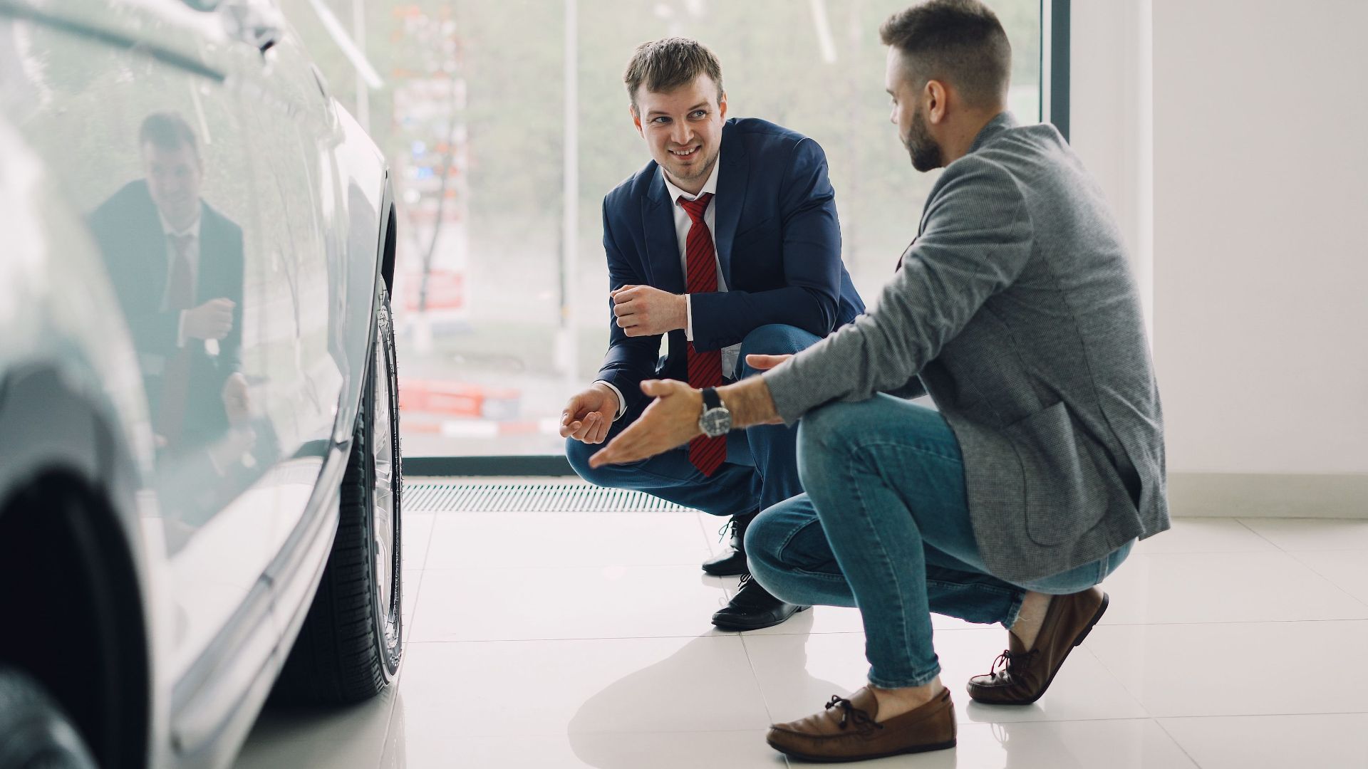 Two men discussing car features in a showroom, kneeling near a vehicle.