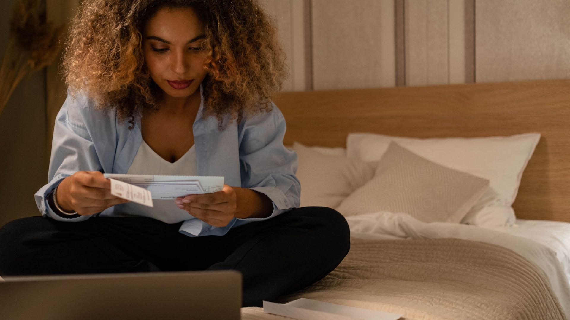 Woman sitting on bed reading documents with laptop, highlighting home office setting.