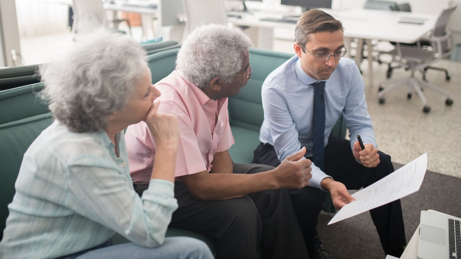 Business professional consults elderly clients in an office setting. Collaborative discussion, paperwork visible.