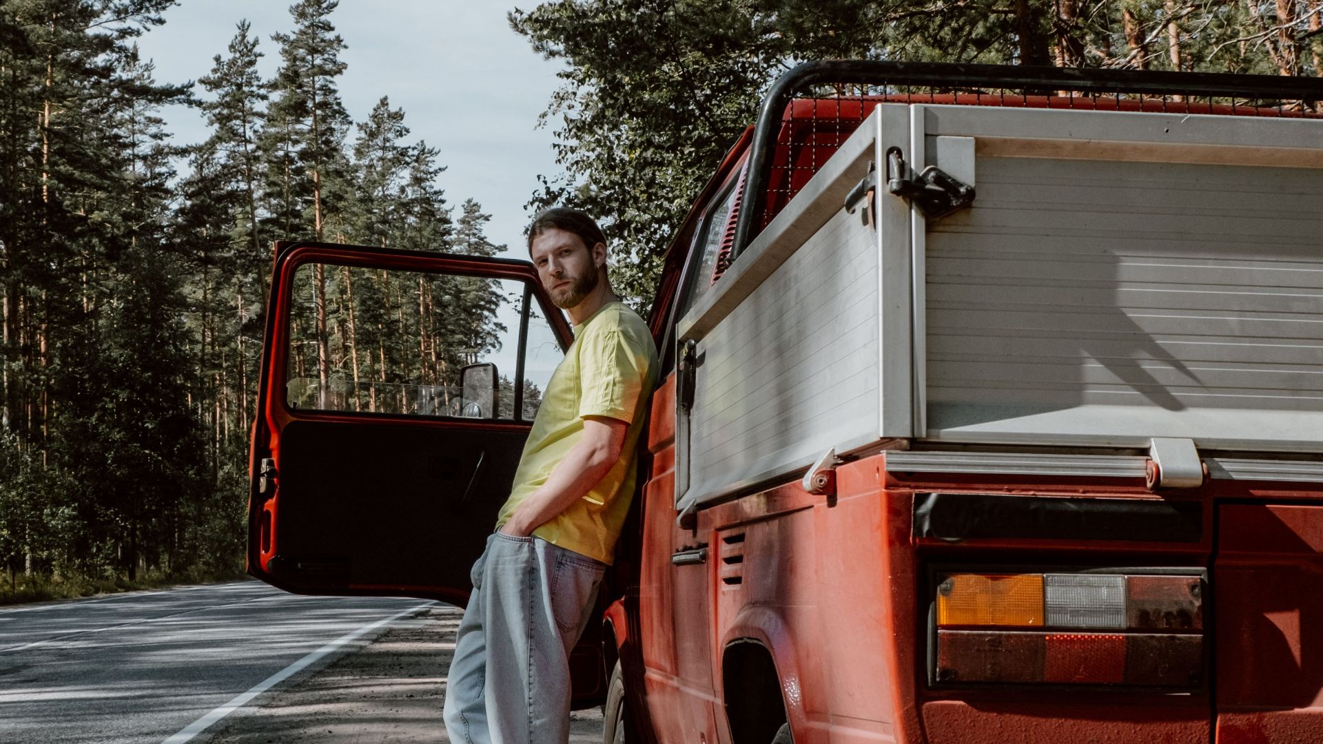 A man stands by his red pickup truck on a quiet country road surrounded by tall trees.