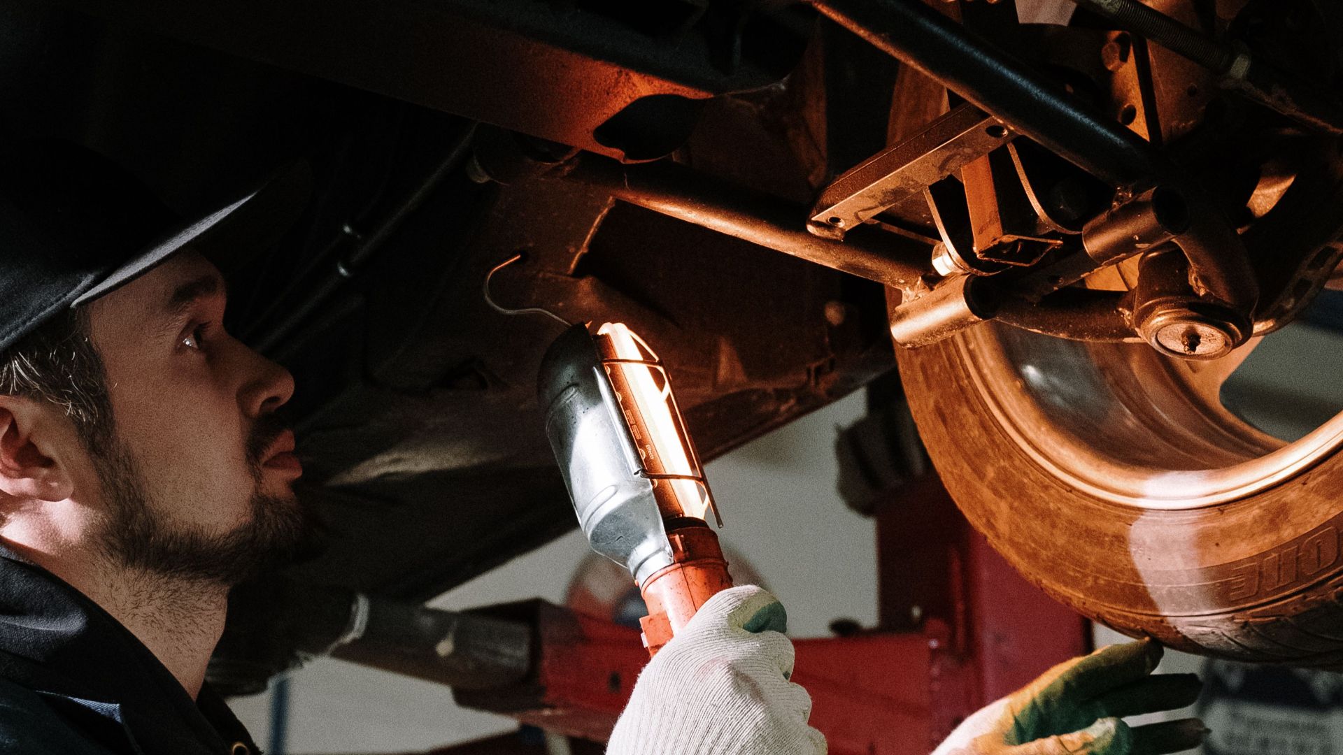 Mechanic uses a flashlight to inspect car's suspension in a garage setting.