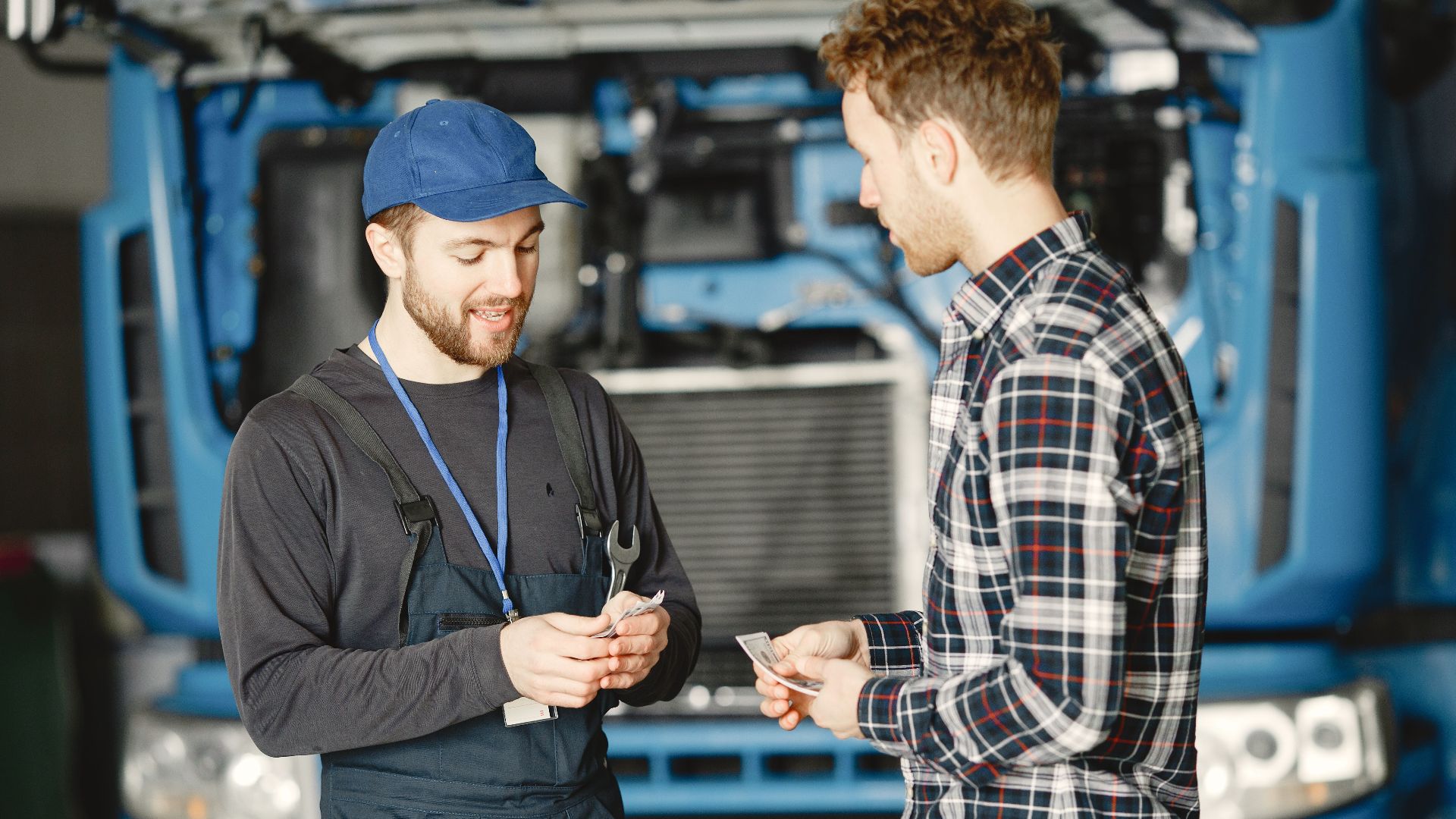 Auto mechanic in blue uniform receives payment from customer in a garage setting with a truck in the background.