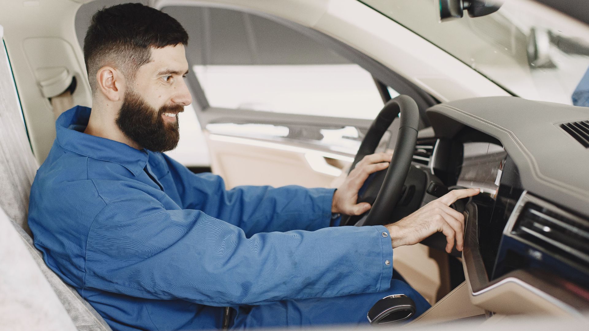Mechanic in blue coveralls interacts with car dashboard, smiling and focused.