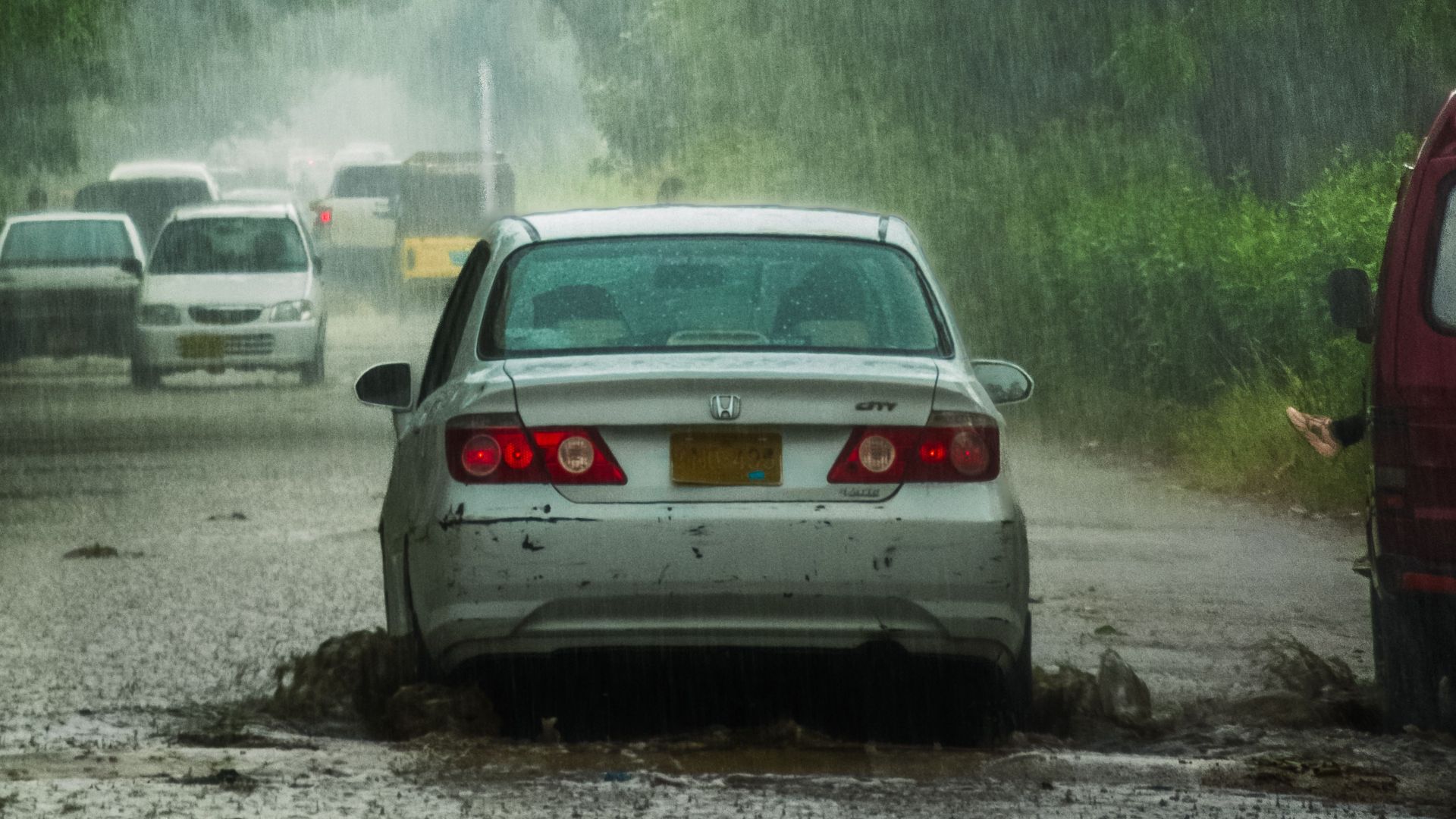 Cars driving through heavy rain on a city street with large puddles and greenery around.