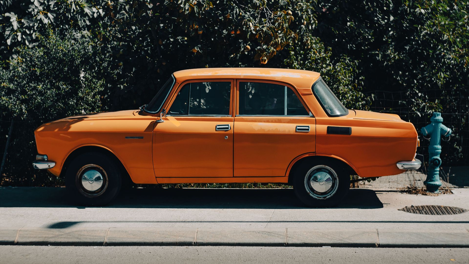 A classic orange Moskvitch 2140 sedan parked on a city sidewalk with lush greenery.
