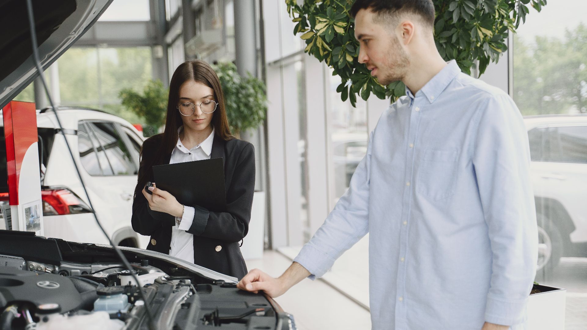 Man and woman examining car engine in dealership service area, focused on vehicle maintenance and functionality.