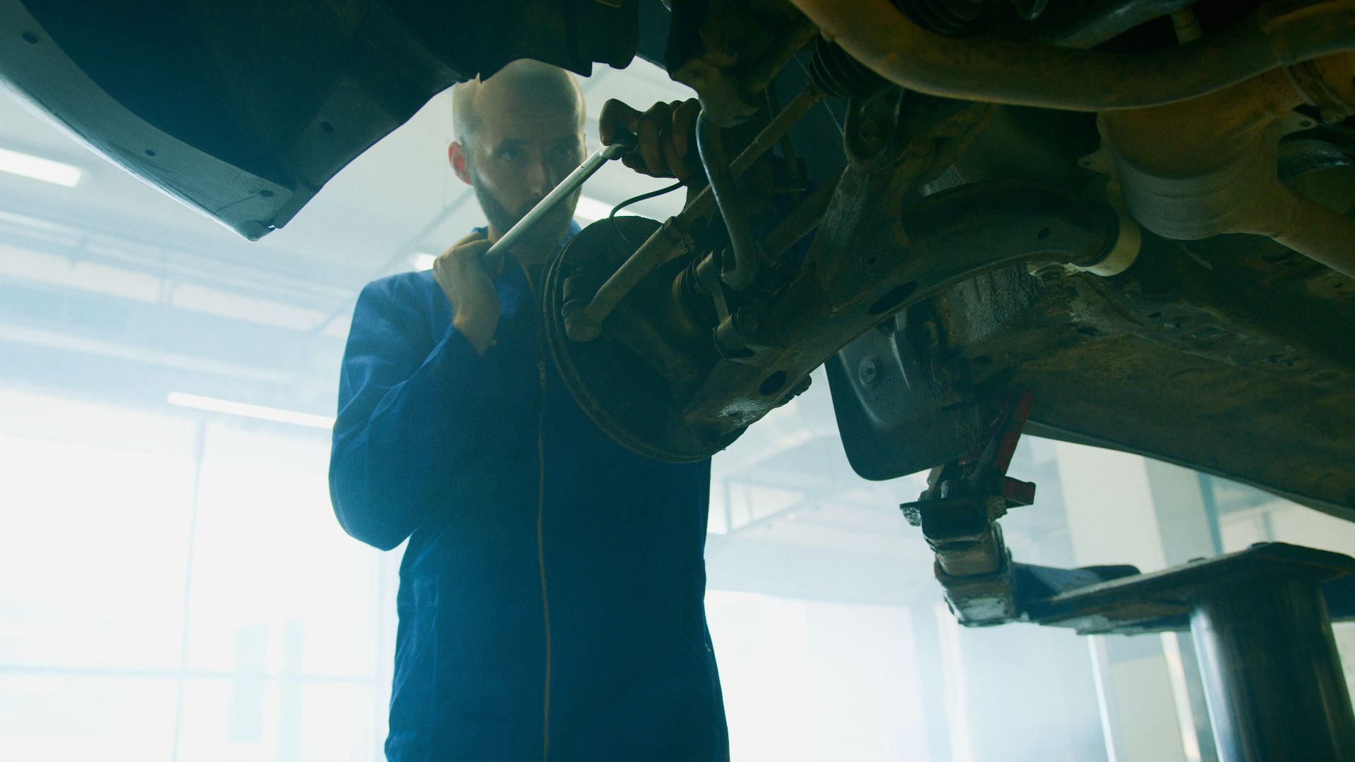 Mechanic in blue coverall fixing a vehicle in a well-lit auto workshop.