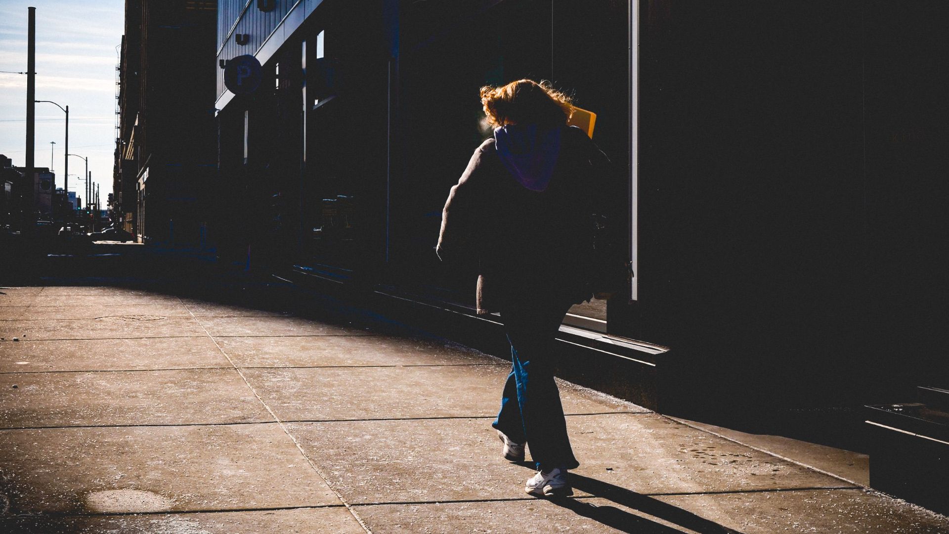 Person walks along a shadowed city street beside modern architecture highlighting urban solitude.