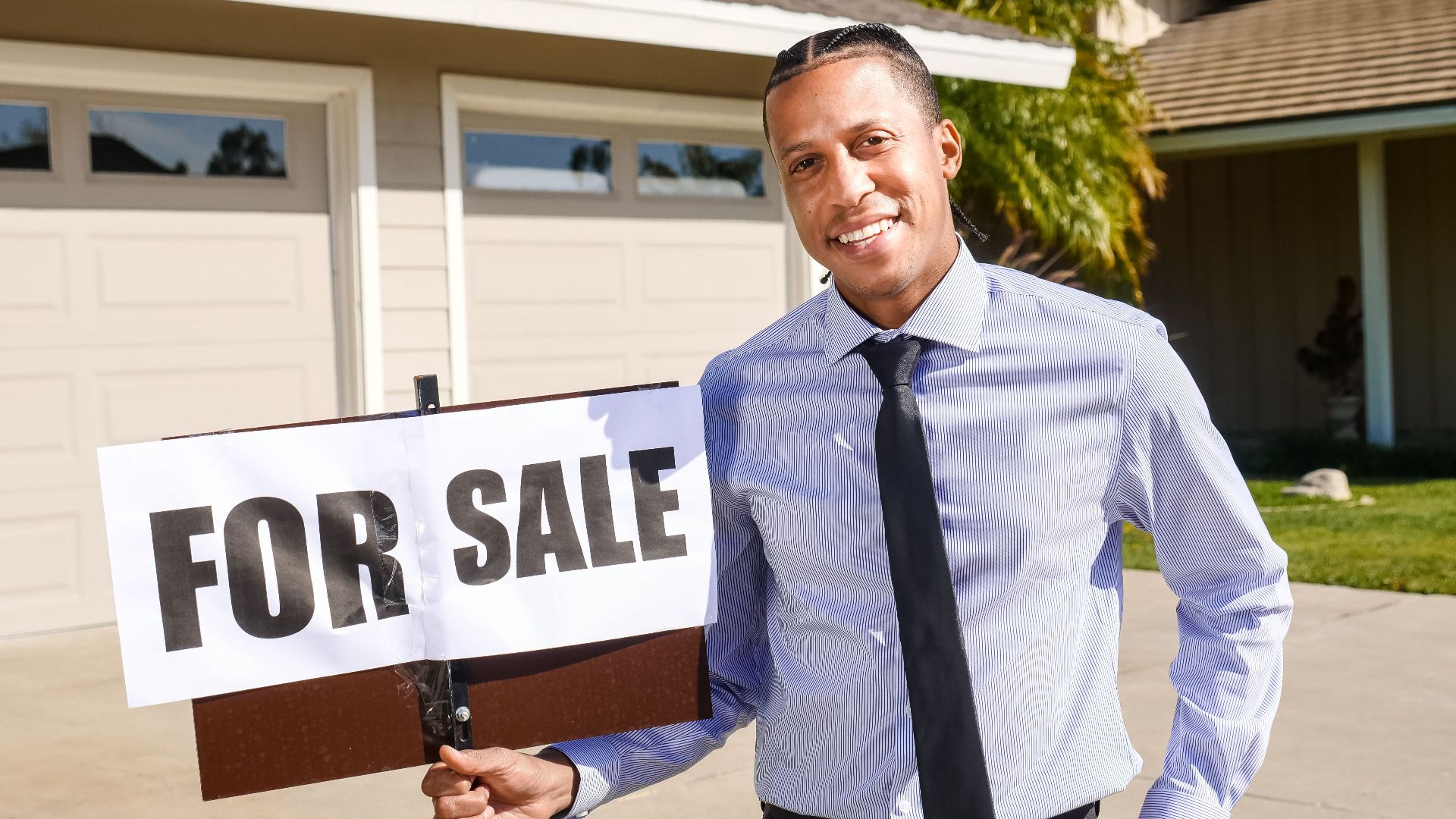 Real estate agent standing with a 'For Sale' sign in front of a house on a sunny day.