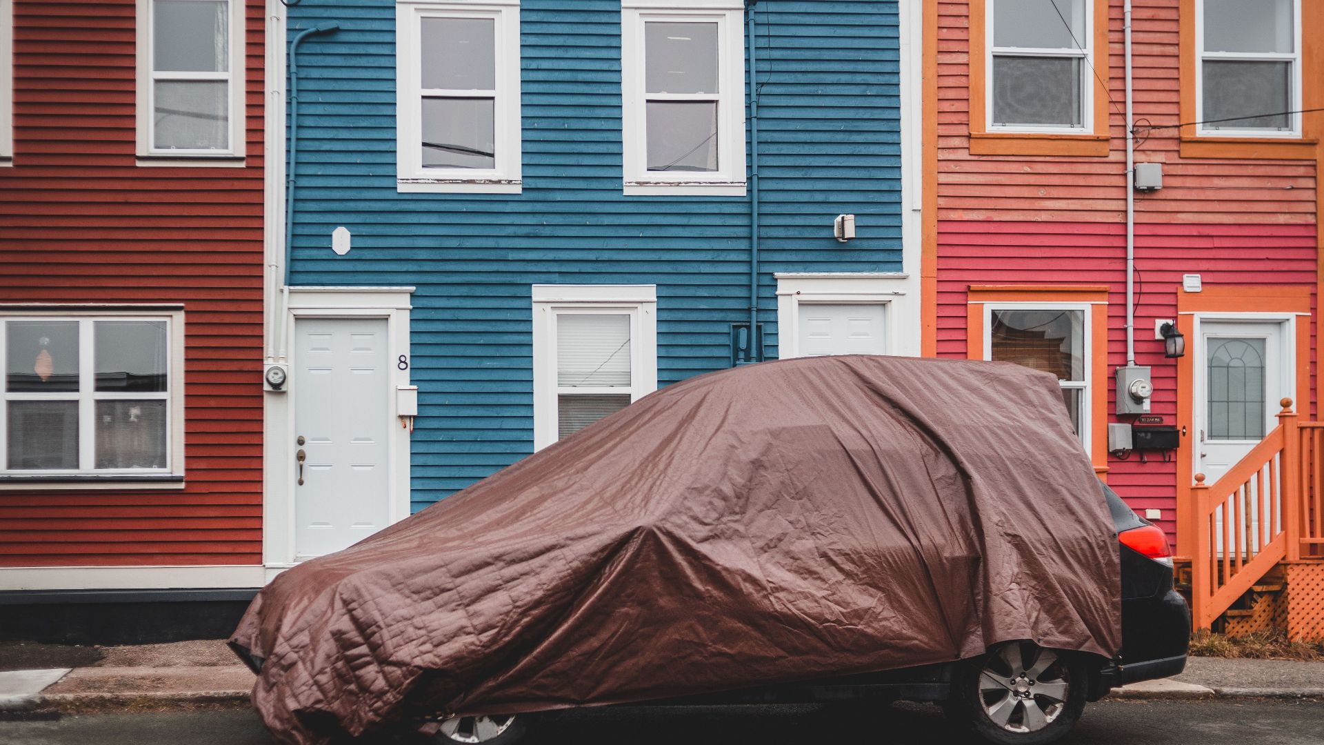 Automobile covered with brown fabric parked on road near colorful residential houses with windows and doors on street of town