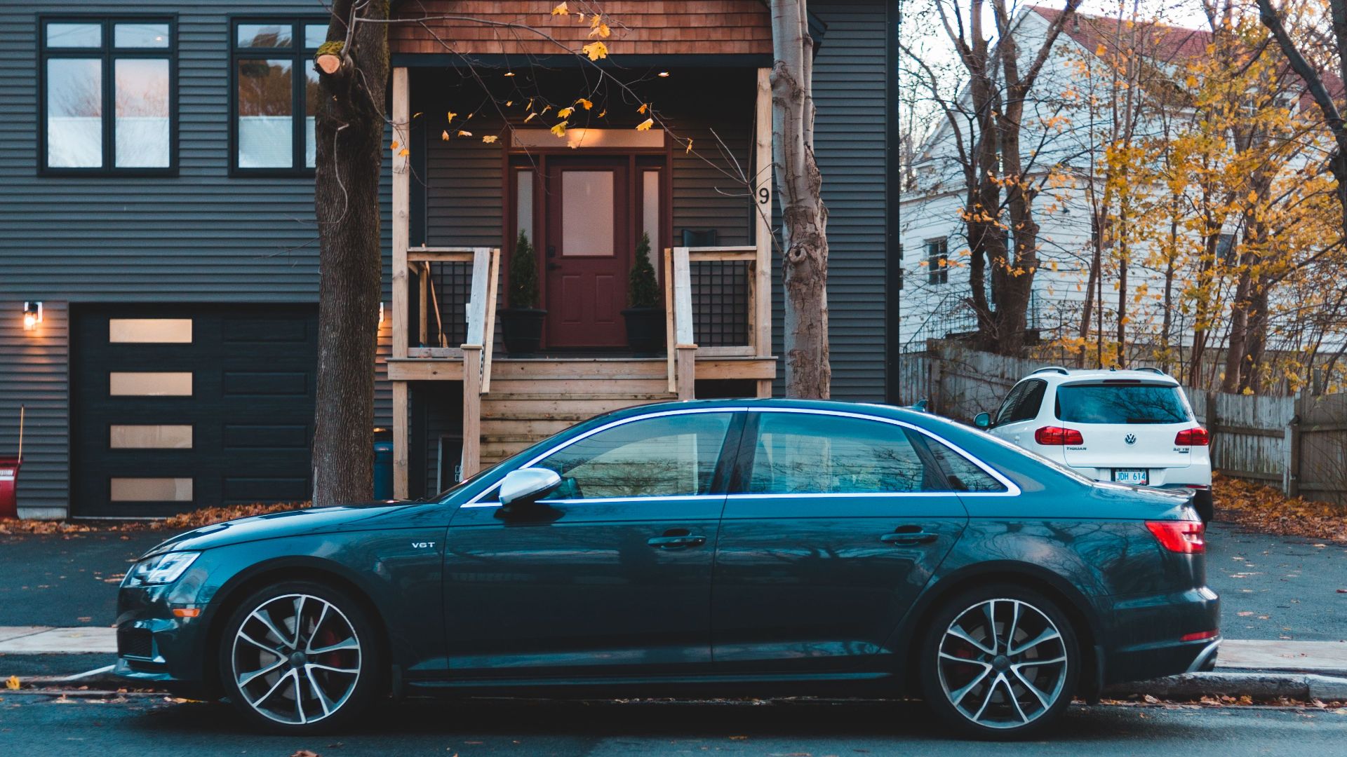 A classic car parked on a street in front of a modern dark-colored house, in an autumn setting.