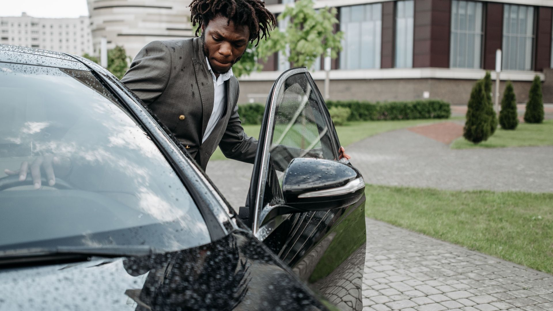An African American man in a gray suit exits a black car in front of a modern office building on a rainy day.