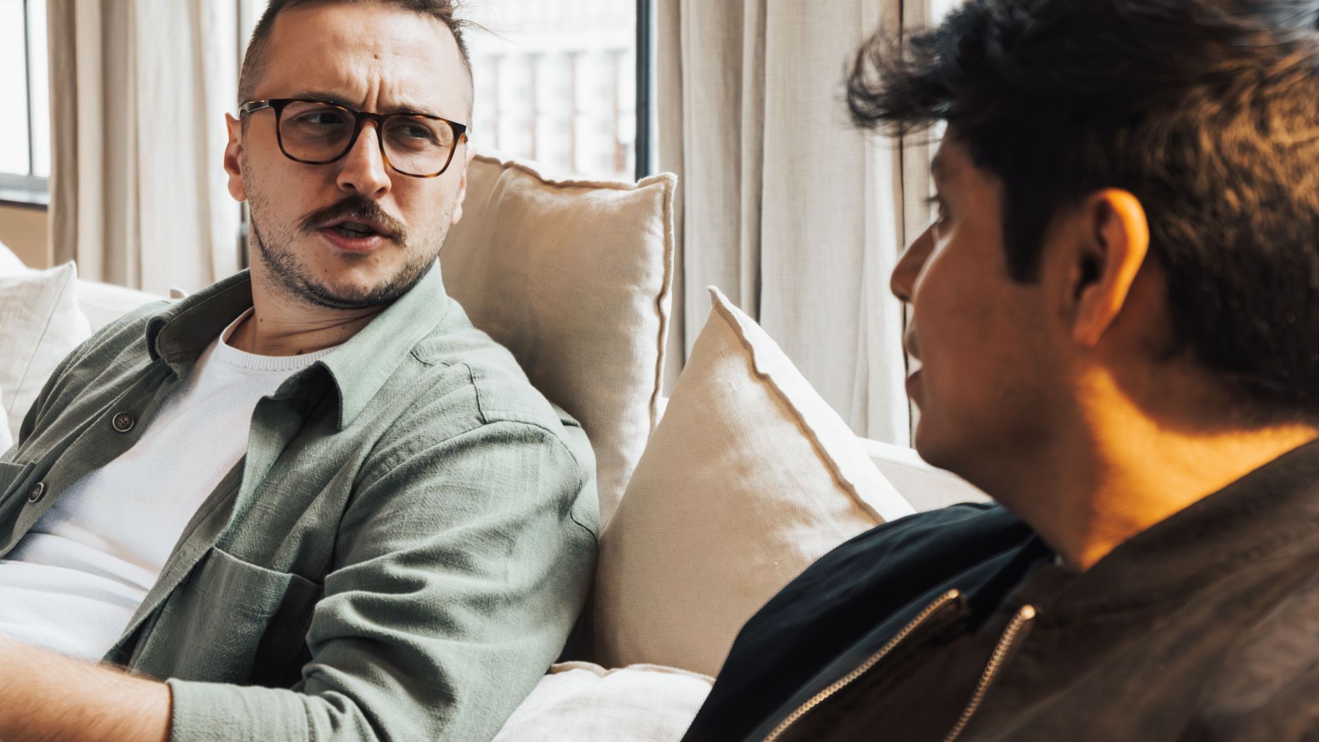 Two men sitting on a sofa, engaging in a conversation while enjoying a coffee indoors.
