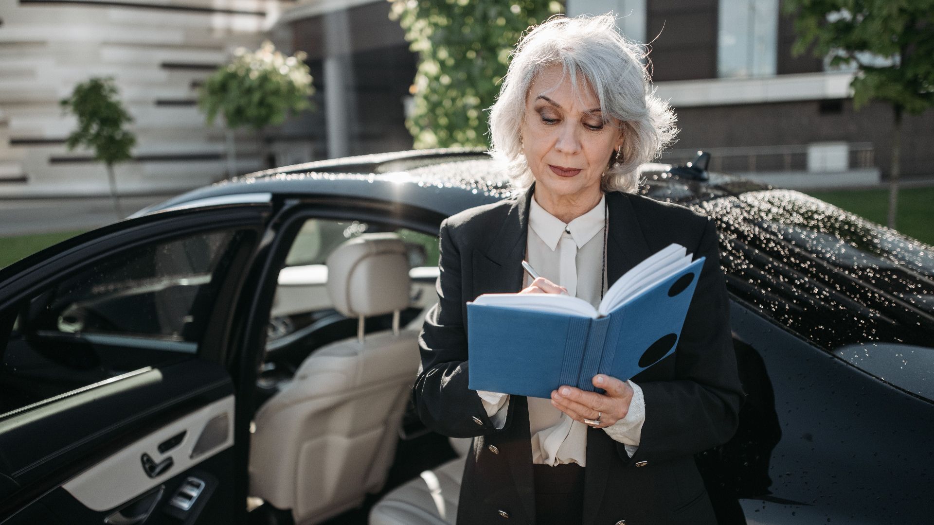 Senior businesswoman writing in notebook next to a car outside a modern building.