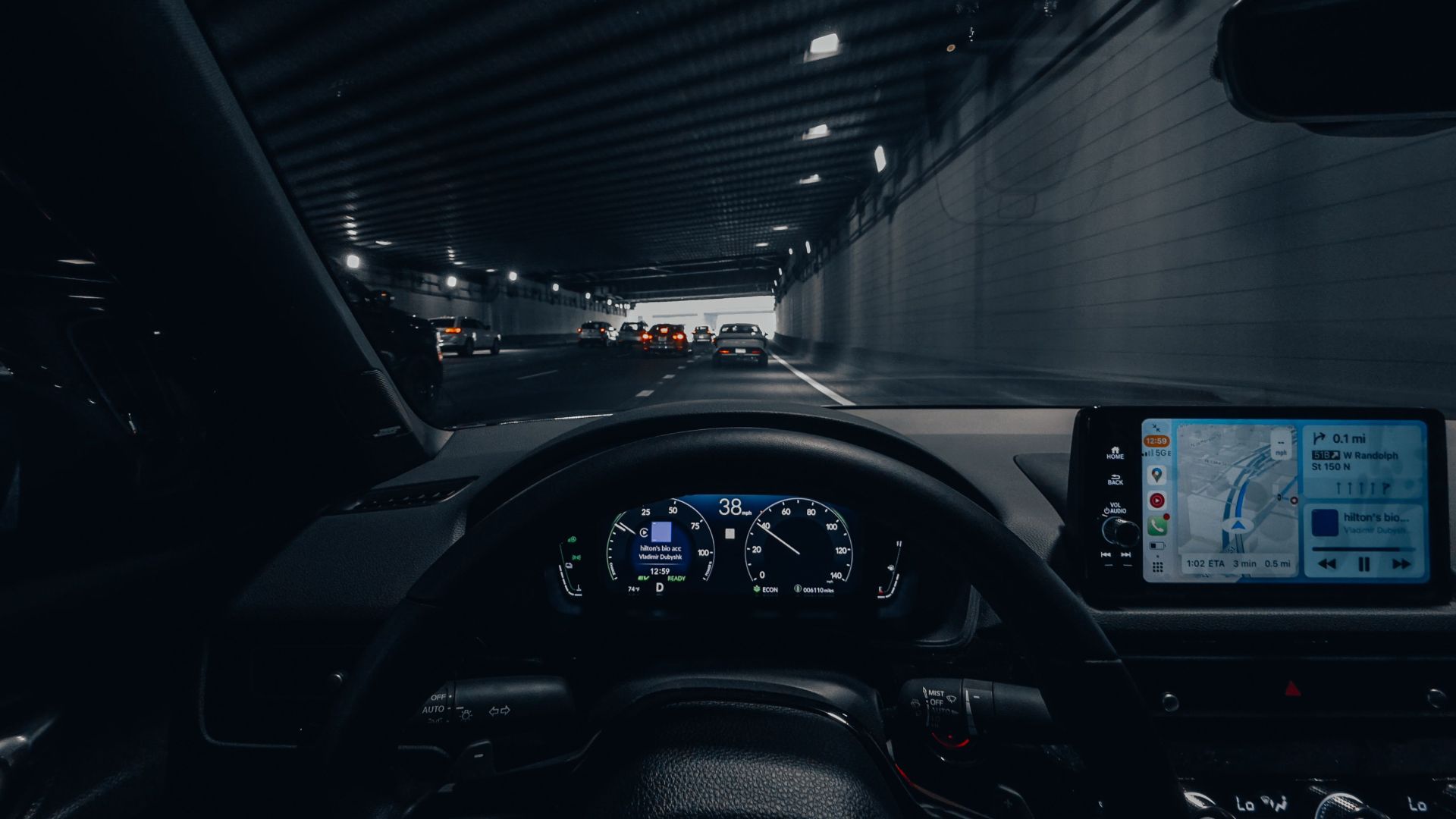 View from car dashboard driving through an illuminated city tunnel at night.