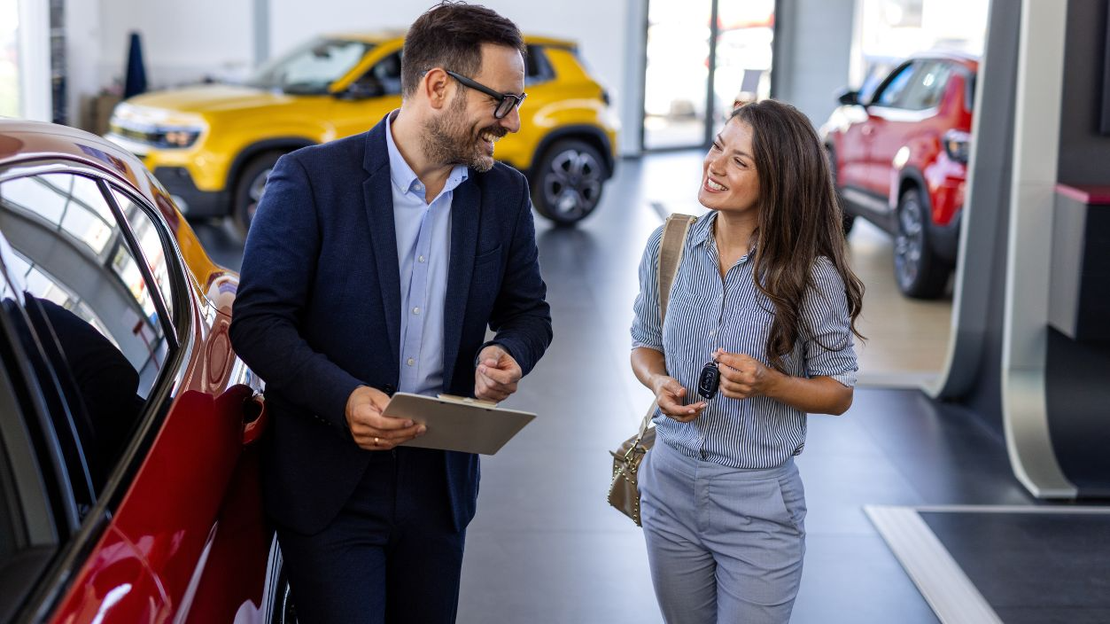 Shutterstock-2532025015, Beautiful young woman is talking to handsome car dealership worker while choosing a car in dealership