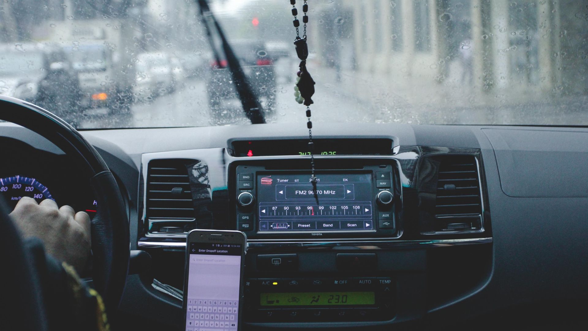 Car interior with driver navigating rainy cityscape using phone and dashboard.