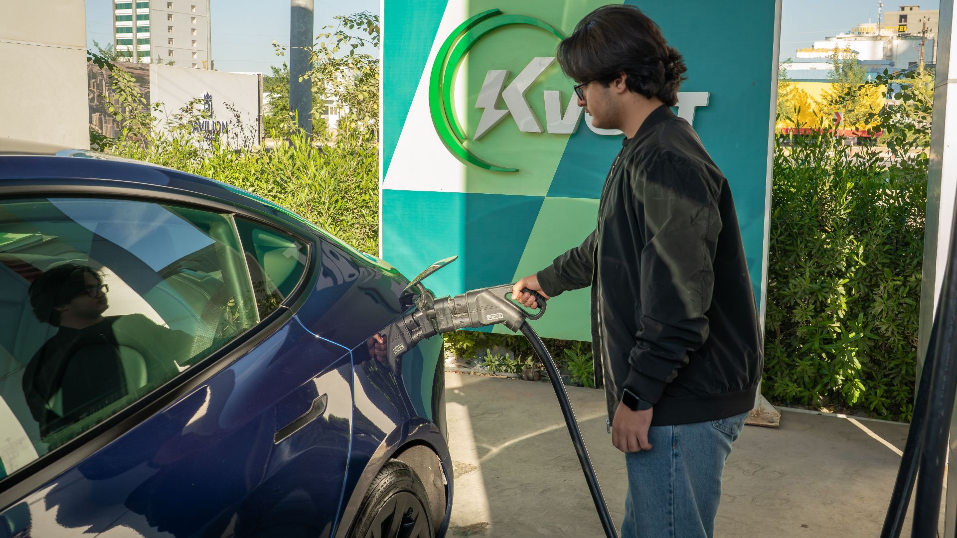 Young man charging a Tesla Model 3 at an outdoor electric vehicle station during the day.