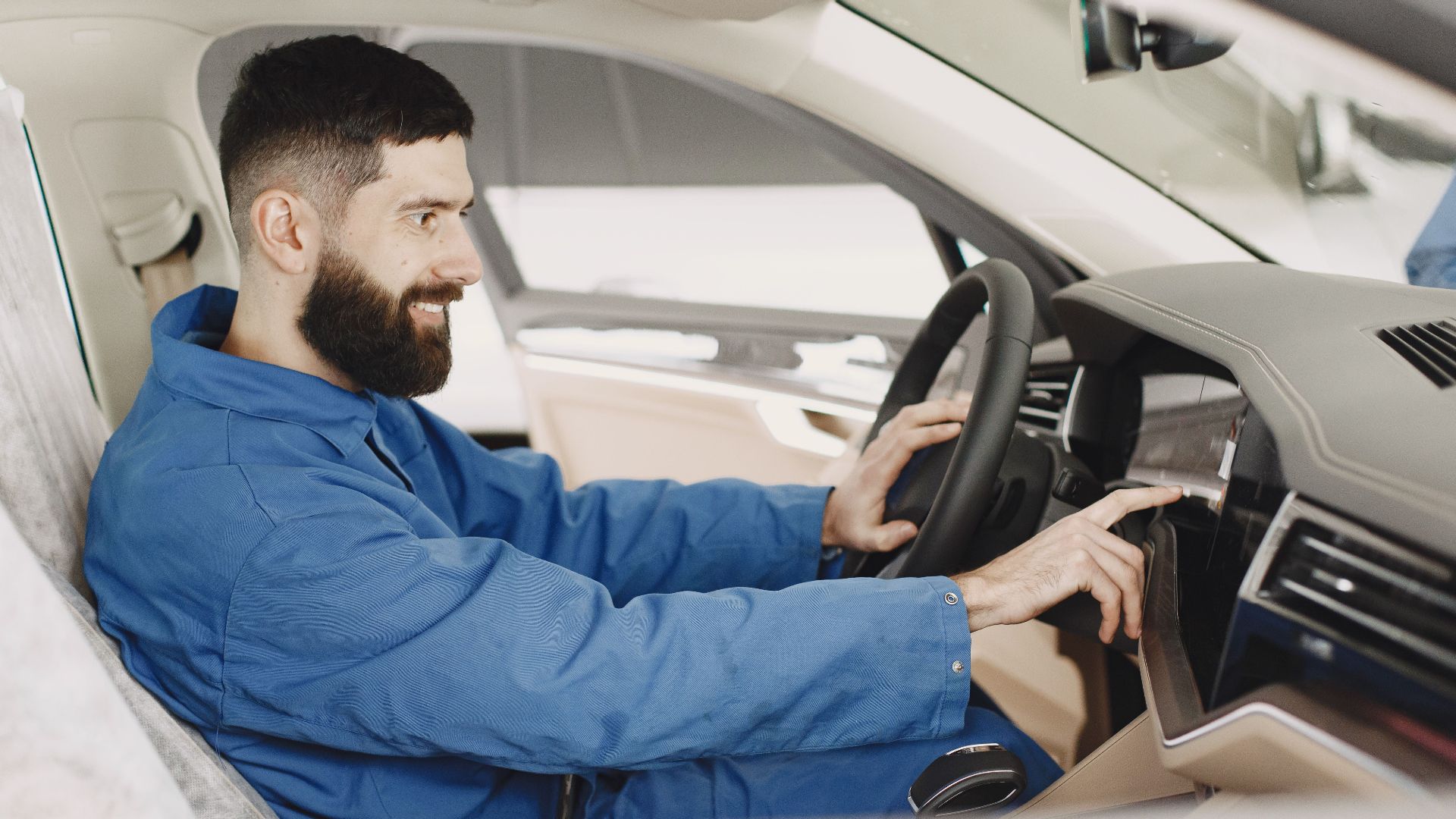 Mechanic in blue coveralls interacts with car dashboard, smiling and focused.