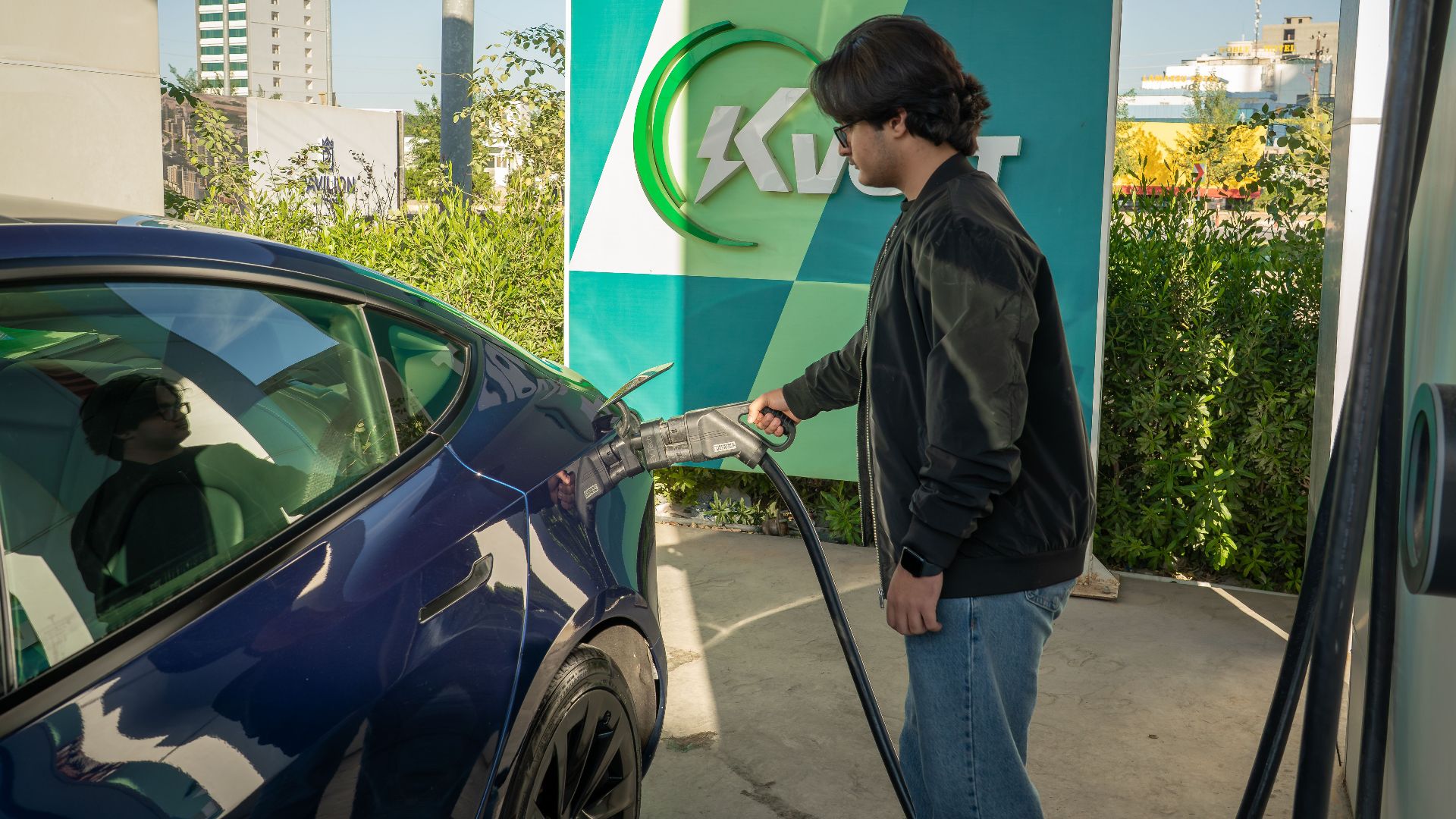 Young man charging a Tesla Model 3 at an outdoor electric vehicle station during the day.