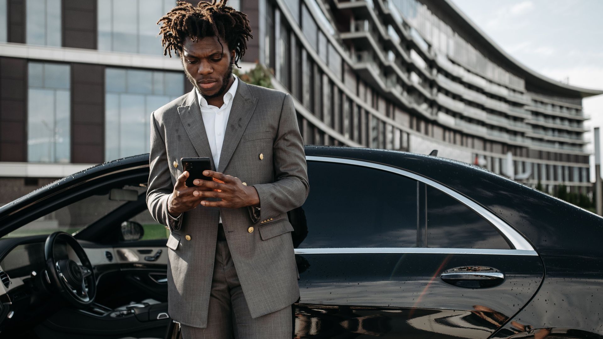 Professional man in a gray suit using his phone next to a luxury car.
