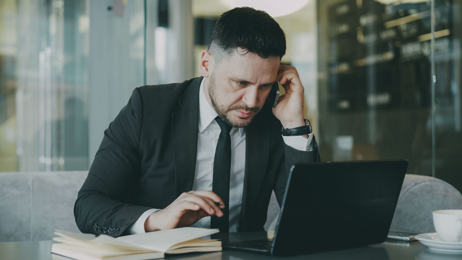 Focused businessman in suit multitasking on phone and laptop in modern office.