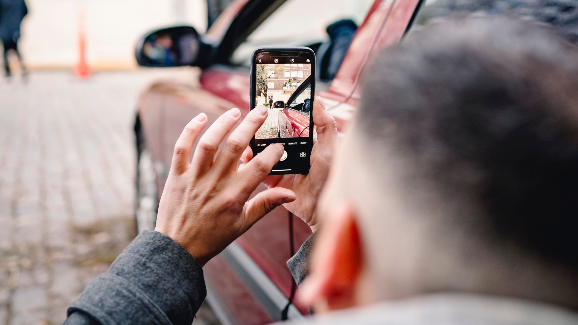 Man using smartphone to take pictures of an urban scene beside a red car.