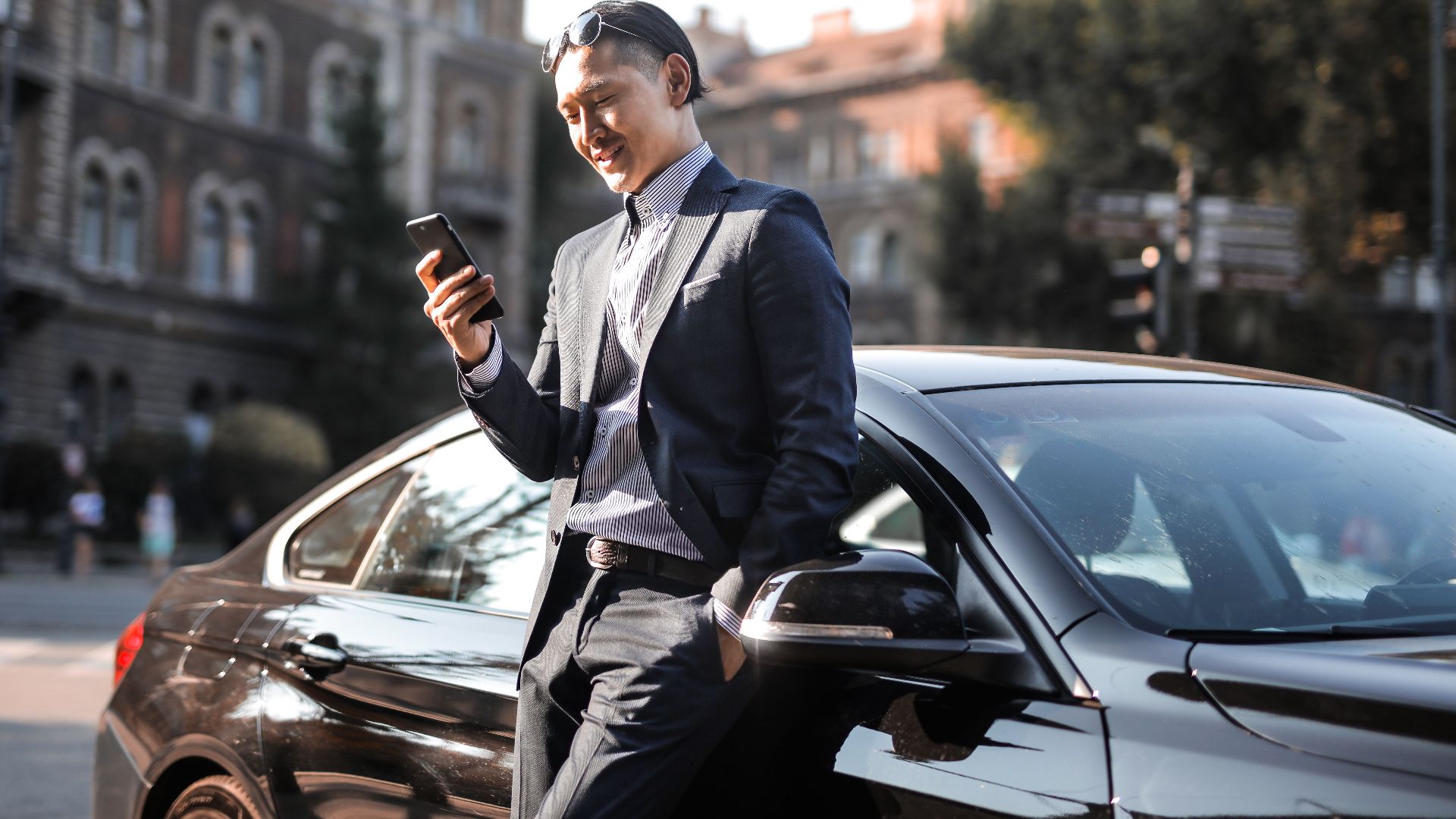 Smiling businessman in a black suit texting on his phone while leaning against a parked car in the city.