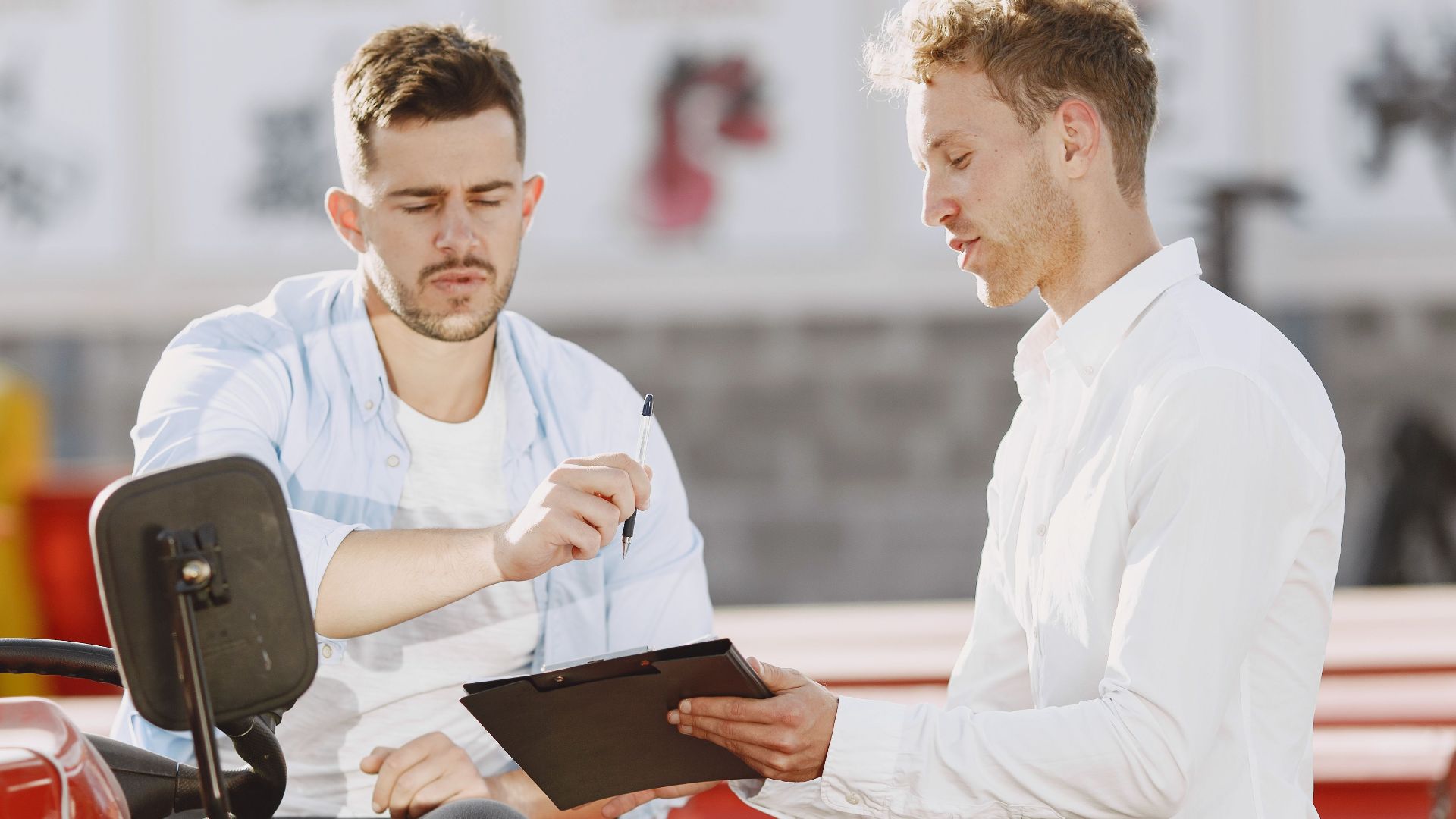 Two professionals discuss on a red tractor, examining paperwork under bright sunlight.