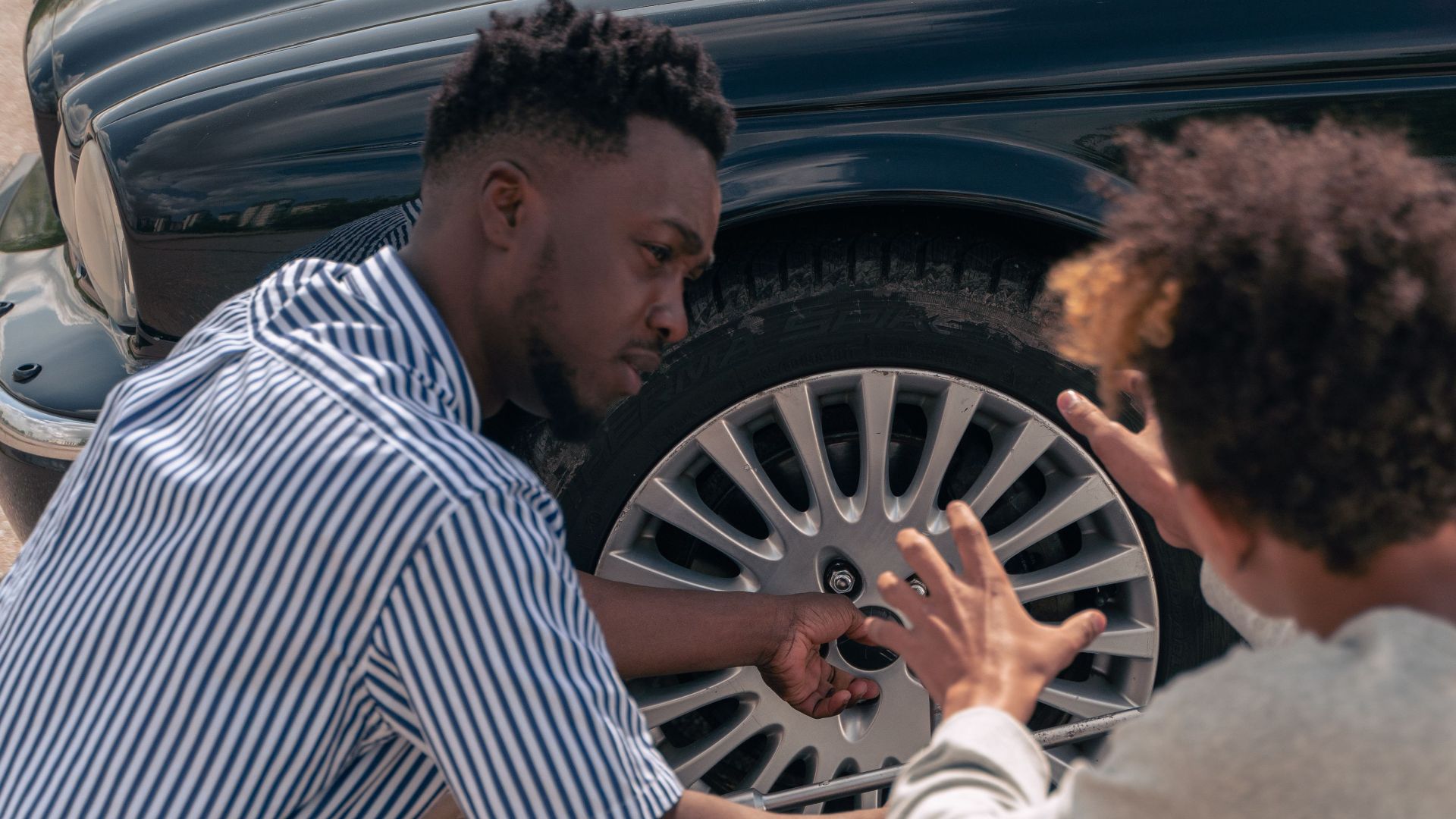 A father instructs his teenage son on how to change a car tire outdoors.