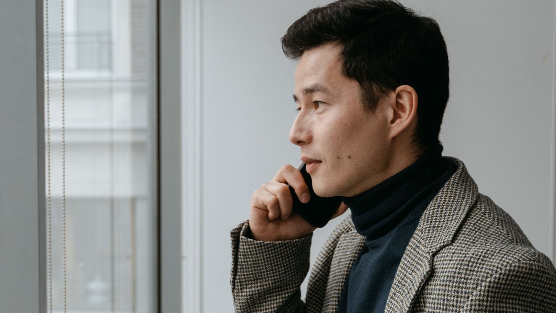 Confident businessman in a gray blazer talking on phone by office window.