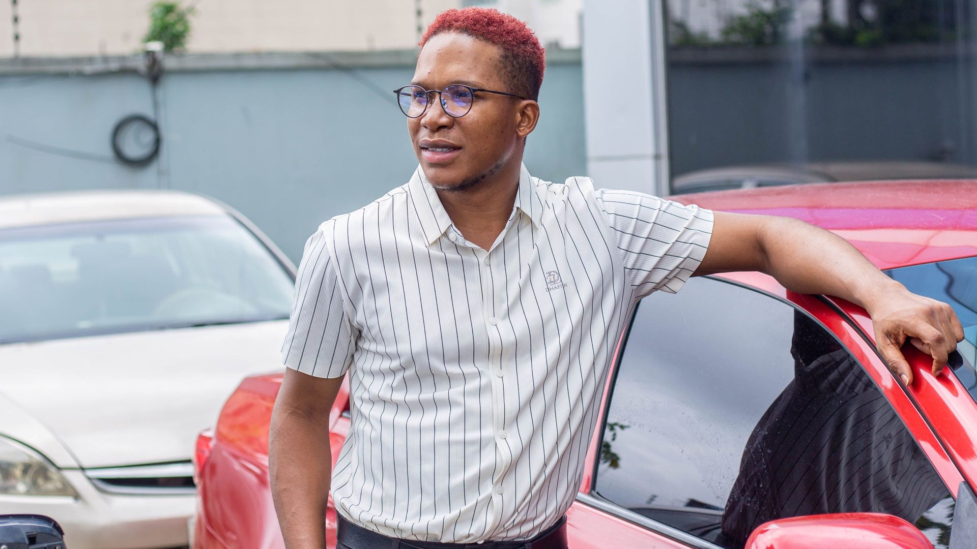 Stylish man wearing eyeglasses poses by a red car in Lagos, Nigeria.