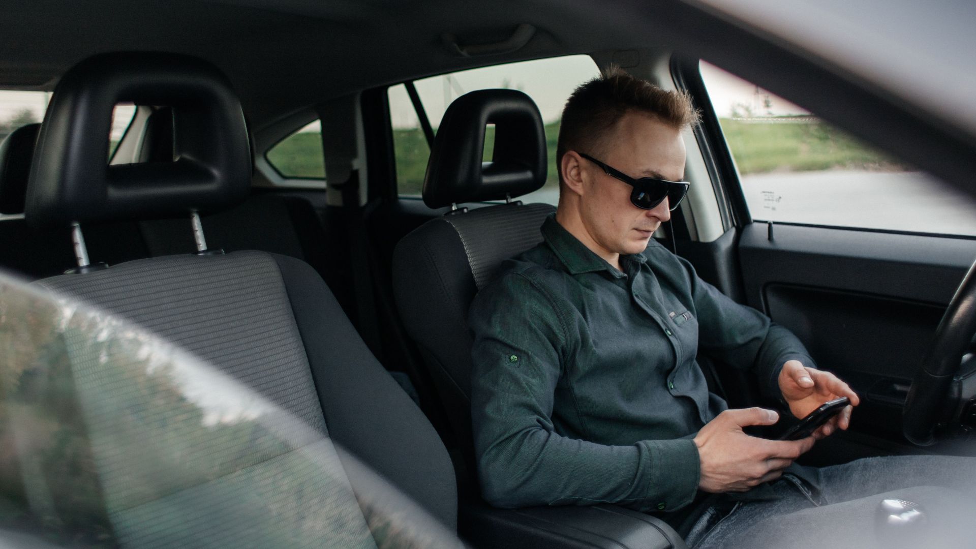 Young man using smartphone inside a parked car, wearing sunglasses.