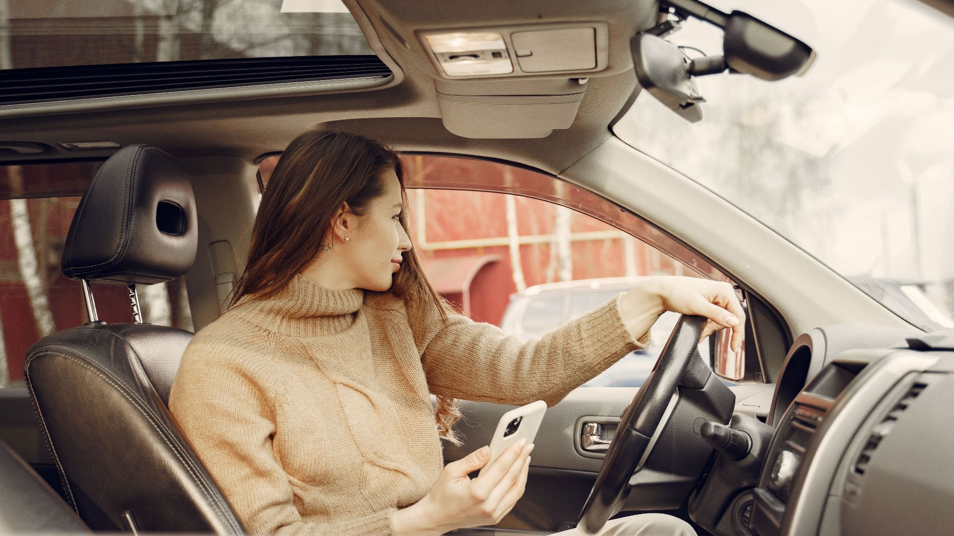 From window side view of adult woman in warm clothes sitting at wheel of modern car waiting for friend and using smartphone