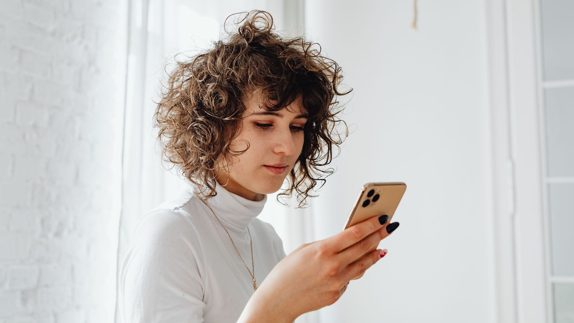 A young woman with curly hair using a smartphone indoors. Natural light and modern decor.