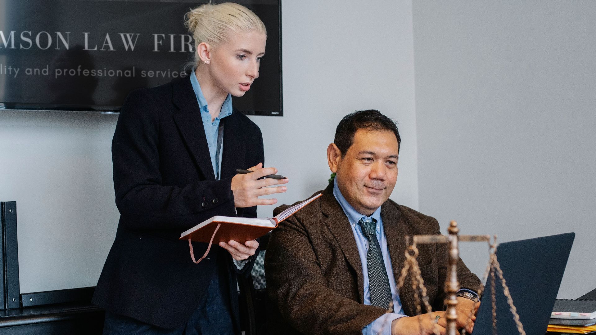 Concentrated young female assistant taking notes in planner while standing at table near middle aged ethnic male colleague typing on laptop during work in law firm