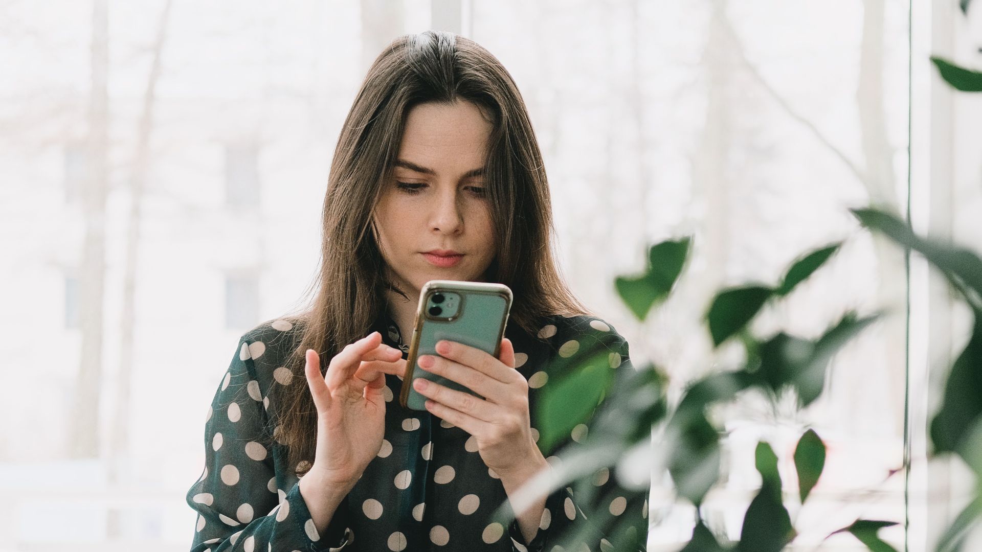 Woman in polka dot dress using smartphone indoors, surrounded by plants.