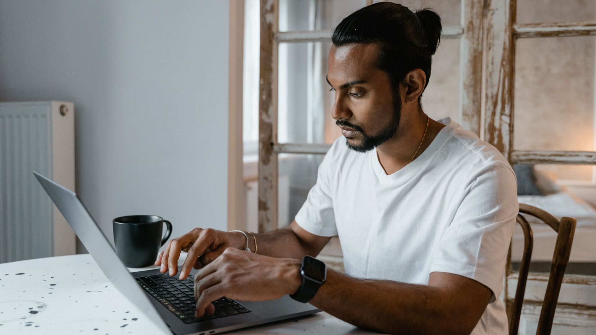 A bearded man is focused while typing on his laptop at home, seated at a desk.