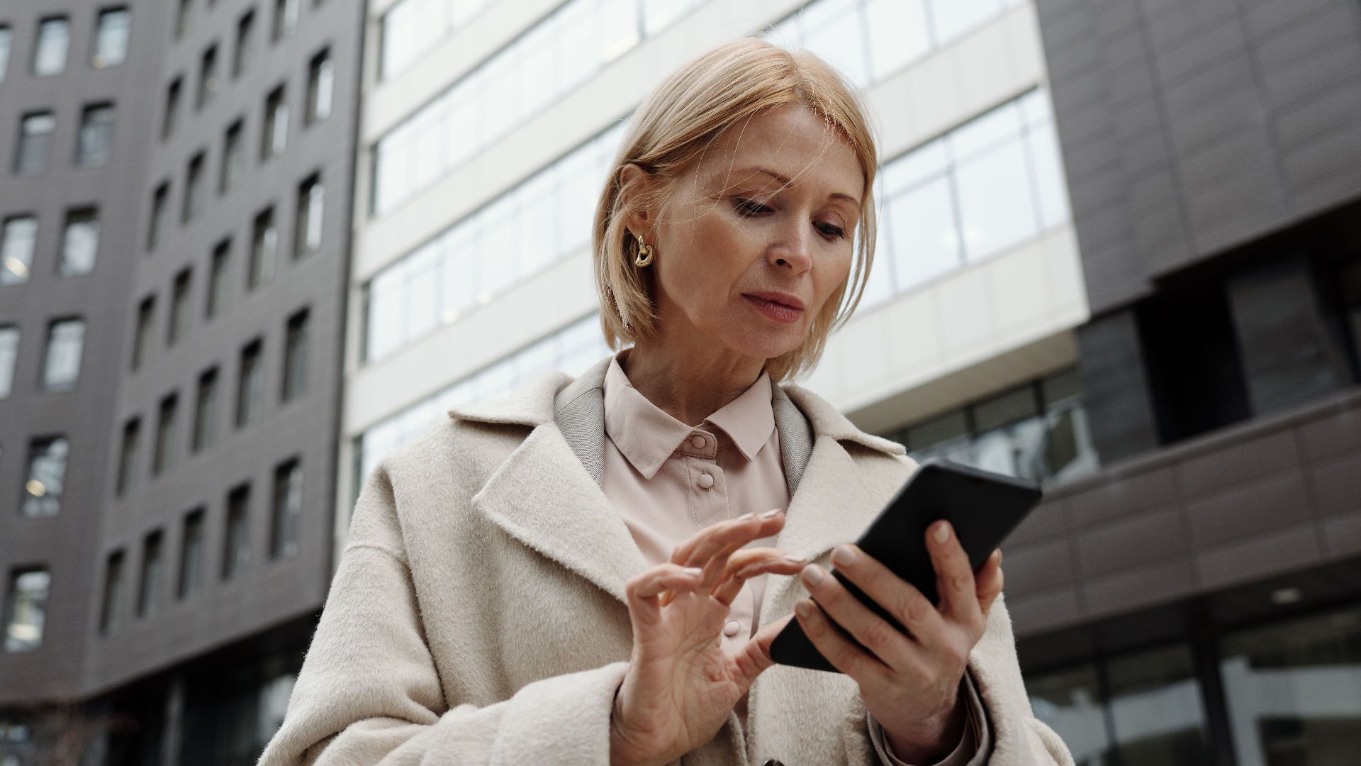 Woman in coat using smartphone in front of modern building. Professional and focused expression.