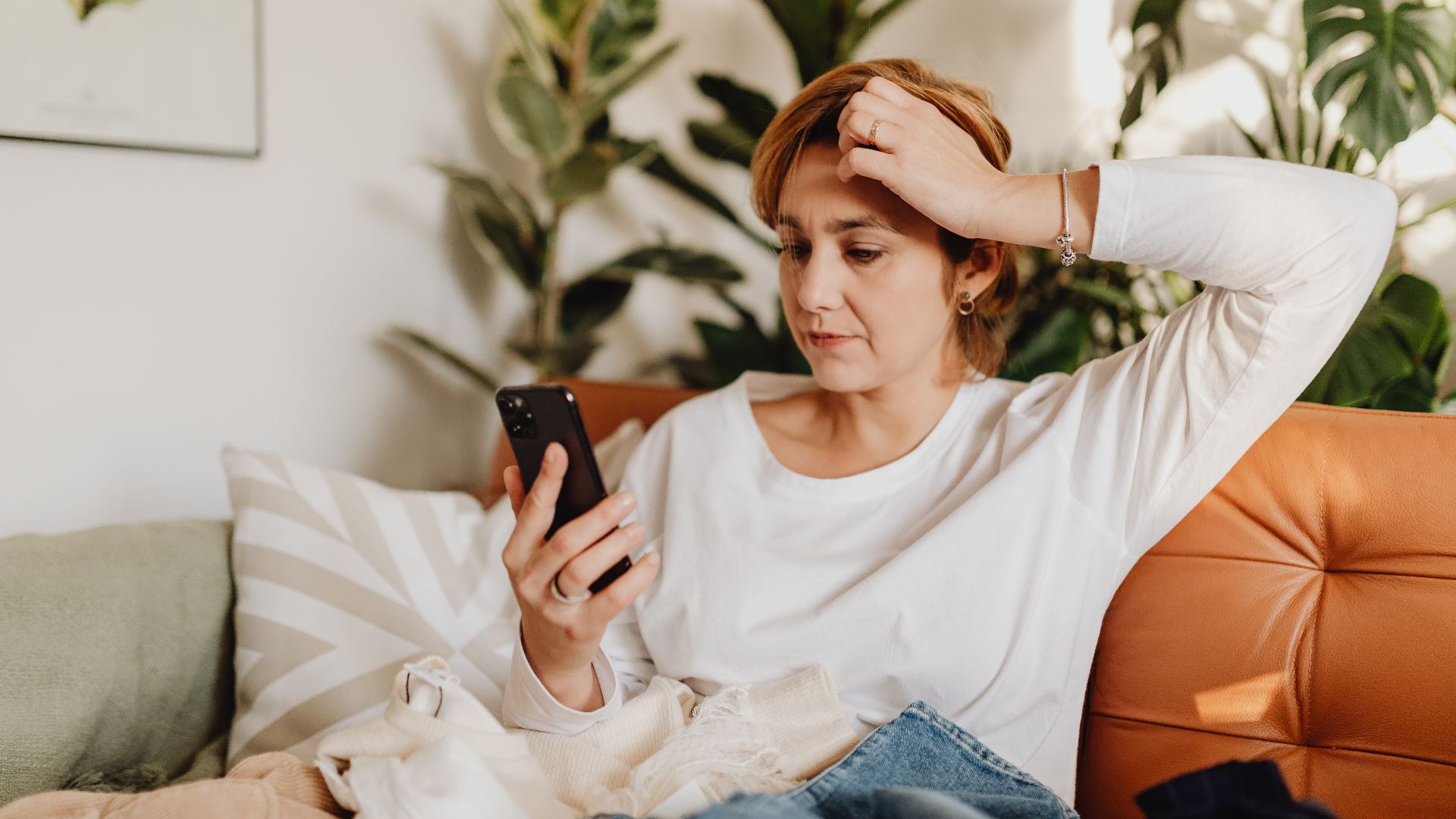 Young woman in white blouse using smartphone while sitting on an orange sofa surrounded by plants.