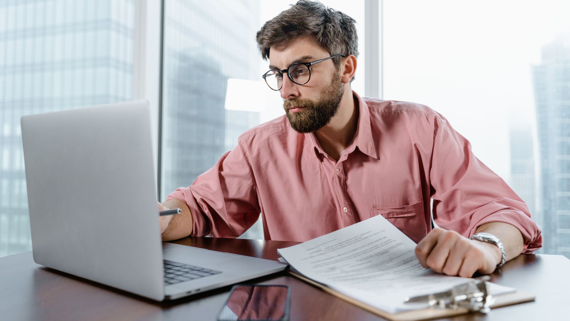 A businessman in an office reviews documents and data on a laptop, concentrating on financial analysis.