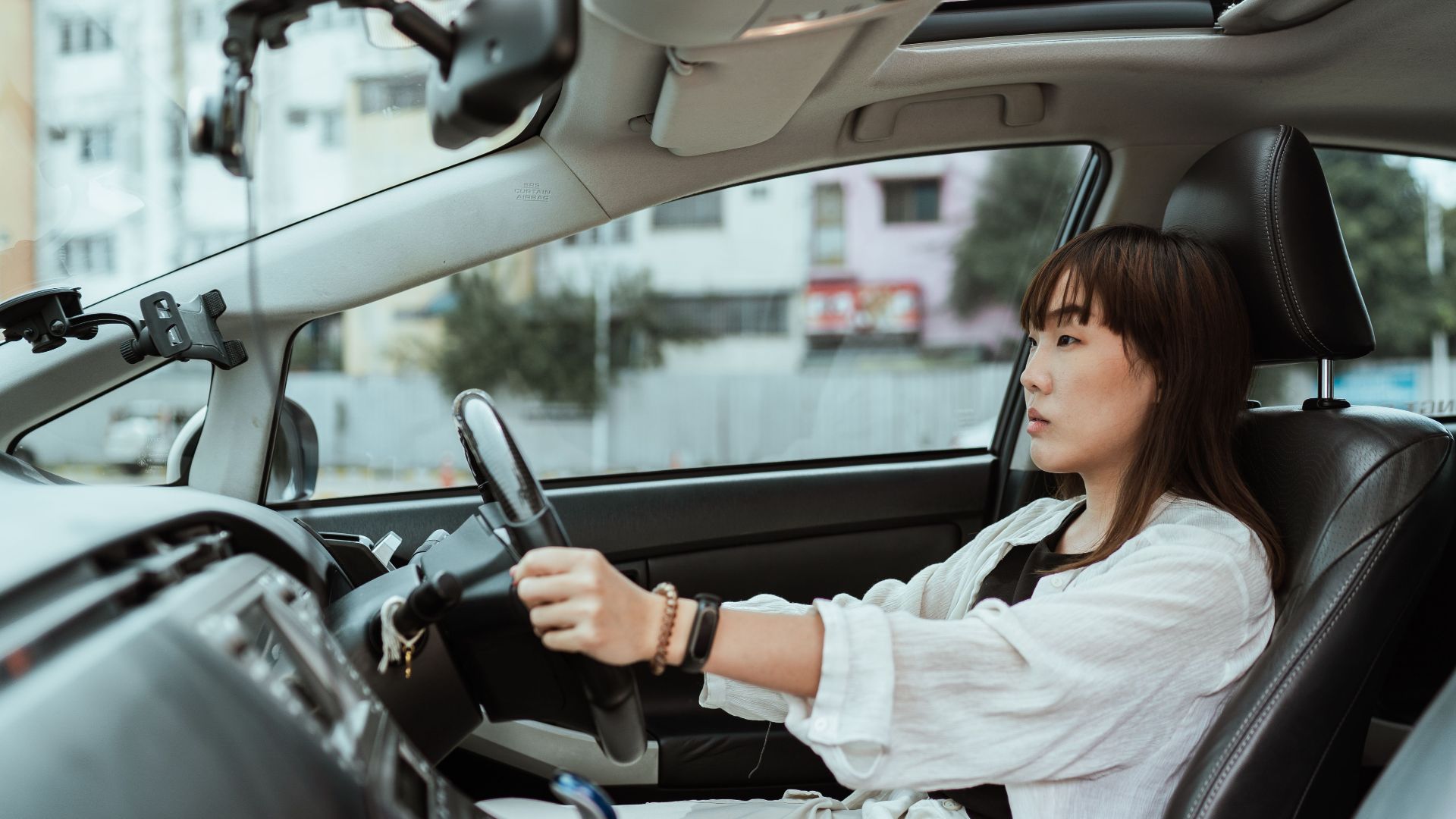 Side view unemotional Asian female in casual white clothes steering right hand drive car along urban streets in daytime