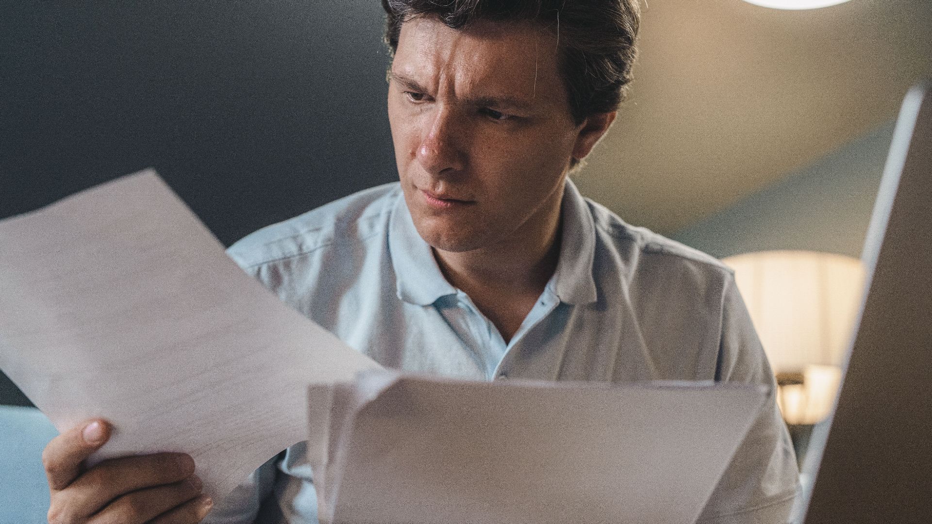 A man intently examines papers, seated indoors under warm lighting, focusing on his work.