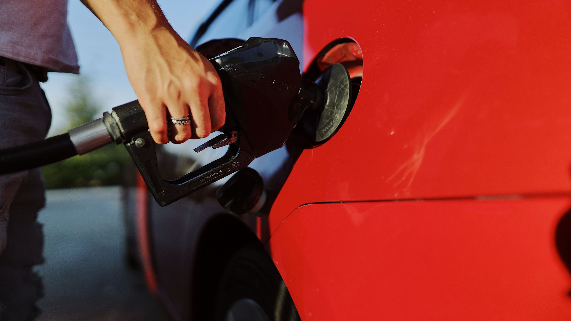 Close-up of a person refueling a red car at an outdoor gas station during the day.
