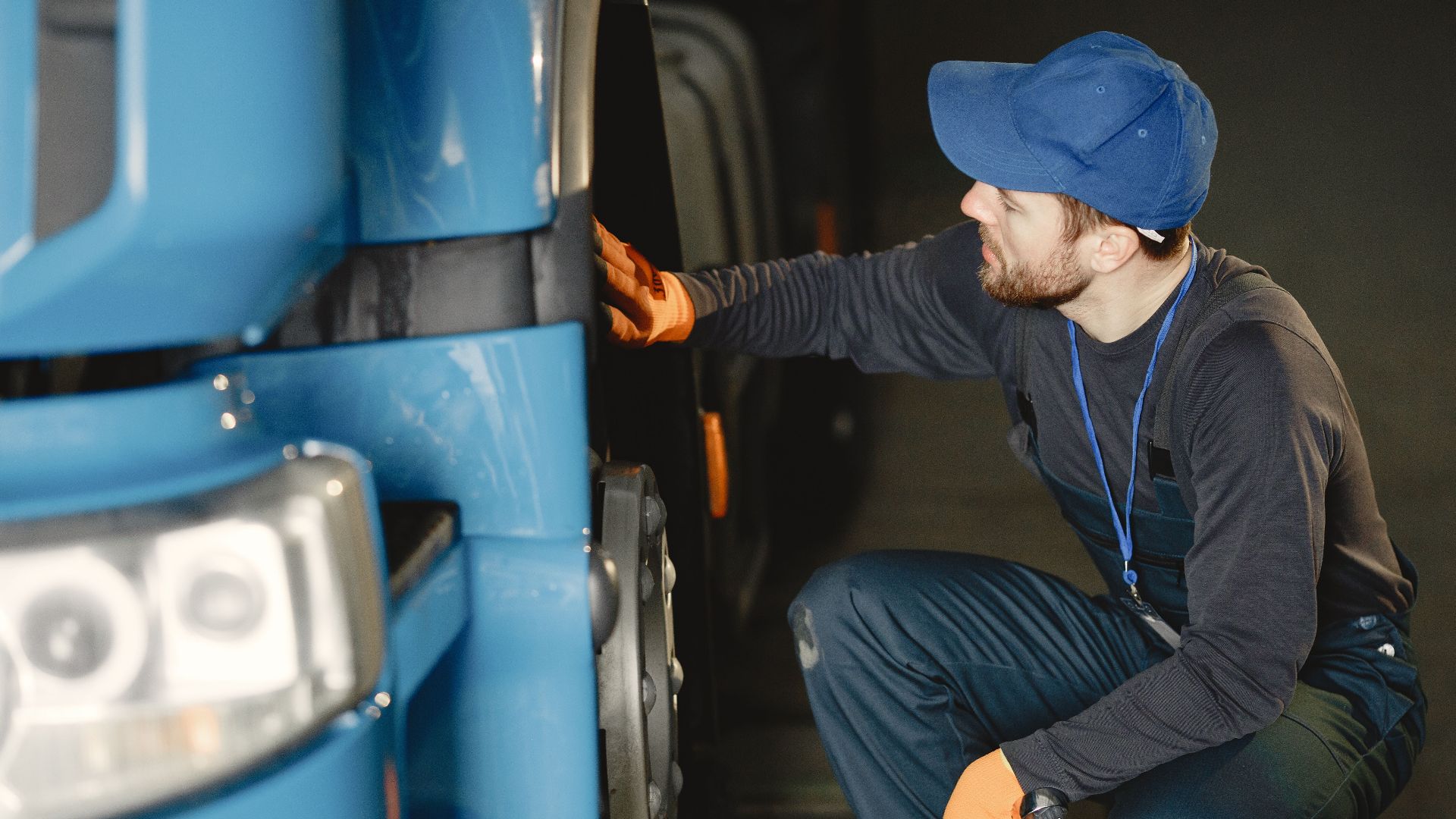 Mechanic in uniform checking a blue truck's wheel inside a garage workshop.