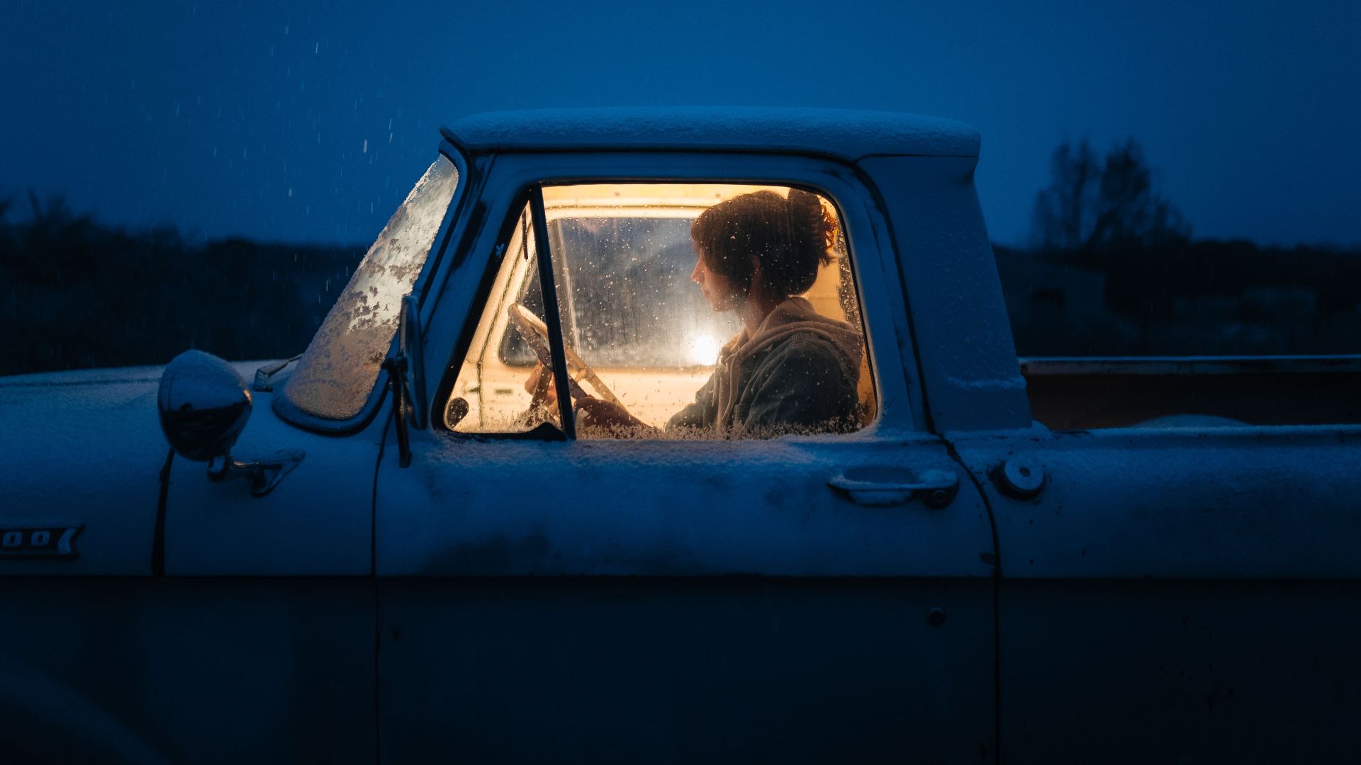 A woman sitting inside a vintage truck covered in snow, illuminated by interior light against a dark winter night.
