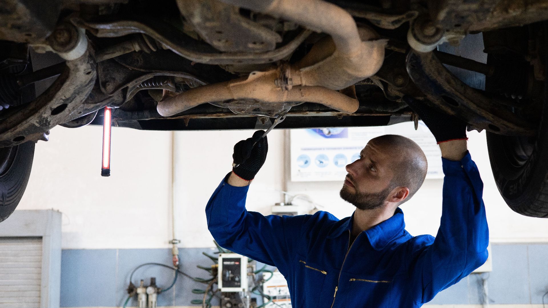 A mechanic in blue overalls inspects the exhaust system of a car in a workshop.