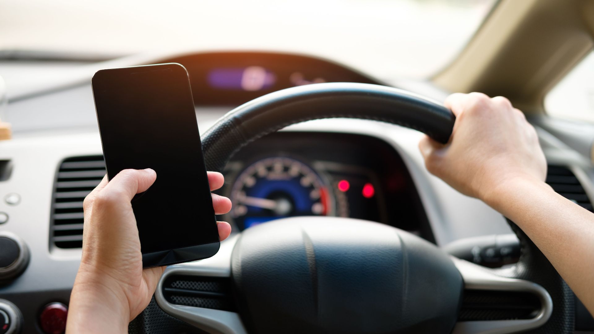A driver uses a smartphone while steering a vehicle, showcasing distracted driving risks.