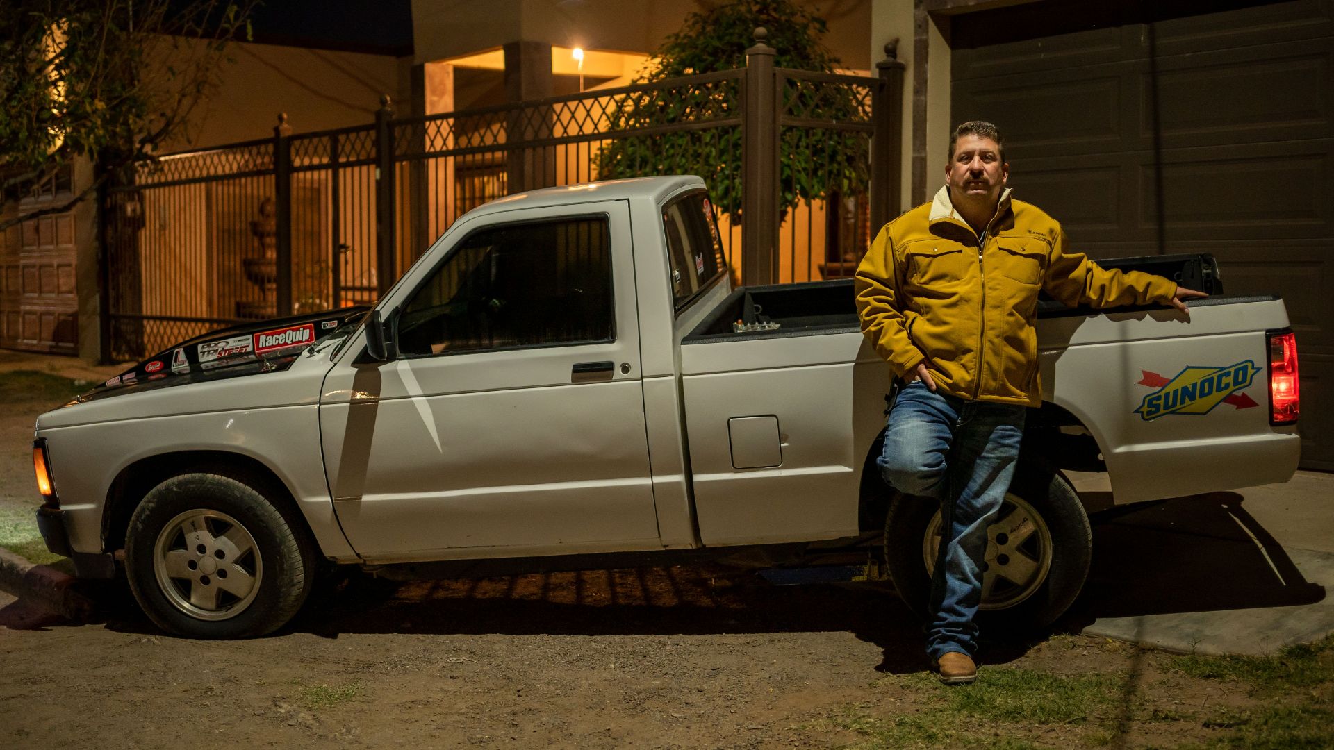 A man in a yellow jacket leaning on a white pickup truck at night in an urban area.